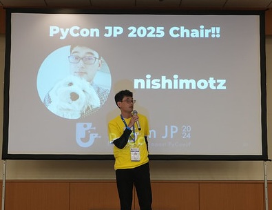 写真 PyCon JP 2025 Chair!! nishimotz と書かれたスライド前で、黄色のスタッフTシャツを着て立っている